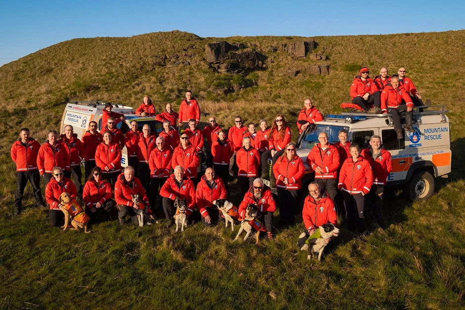 Team members pose for a group photograph. Six search dogs are in the front row and two team vehicles in the background.