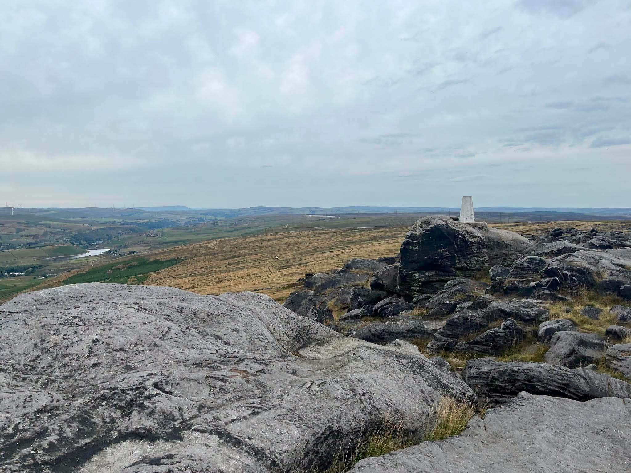 Blackstone Edge trig point stands atop a rocky outcrop overlooking a vast, rolling moorland landscape under a cloudy sky in the English countryside.