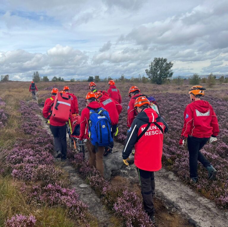 CVSRT in red jackets and orange helmets walk along a path through a field of purple heather, carrying a stretcher.