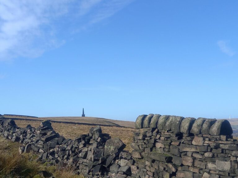 A stone wall runs across a grassy moorland under a clear blue sky, with a tall, slender stone Stoodley Pike monument standing in the distance on the horizon.