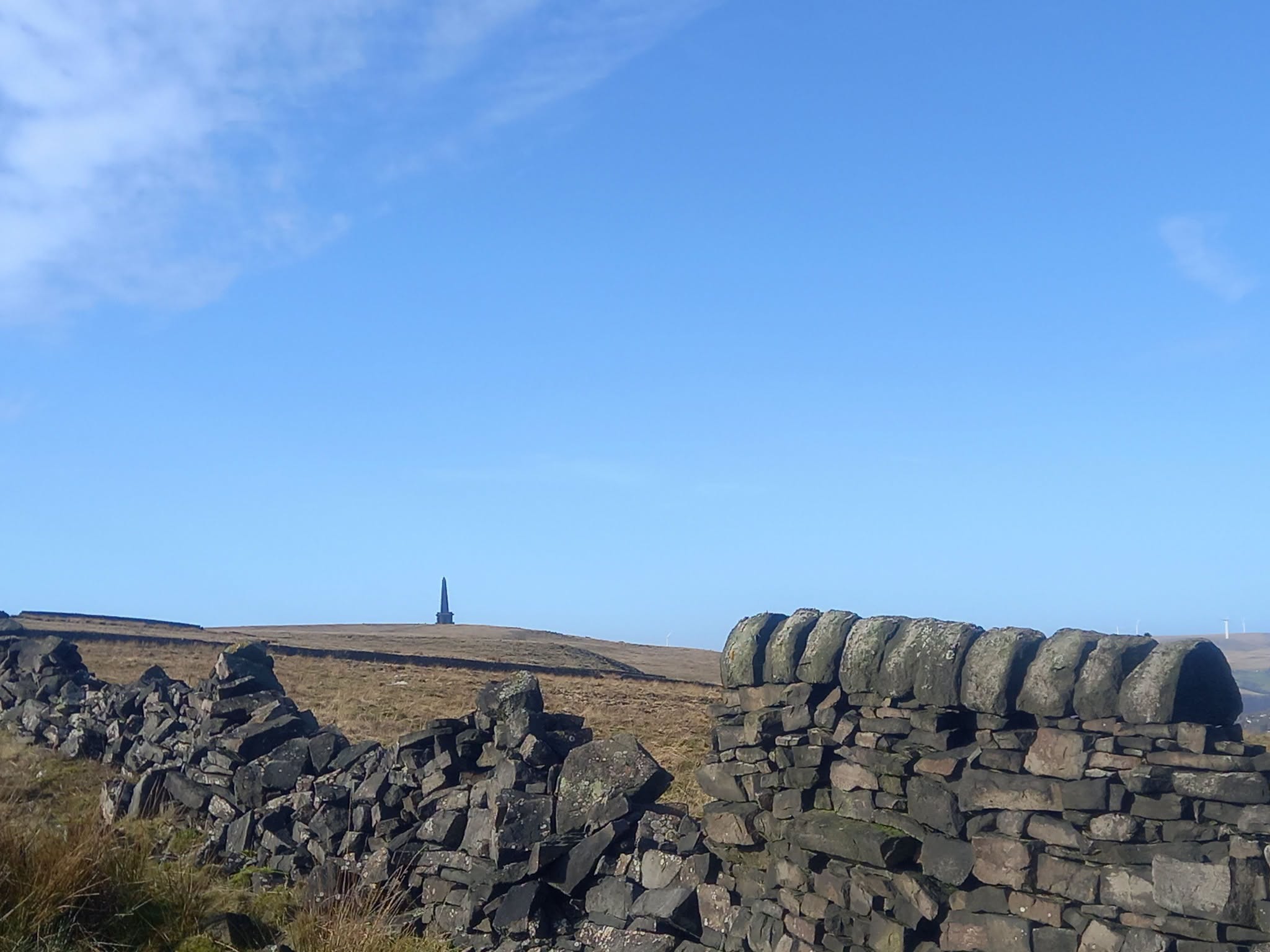 A stone wall runs across a grassy moorland under a clear blue sky, with a tall, slender stone Stoodley Pike monument standing in the distance on the horizon.