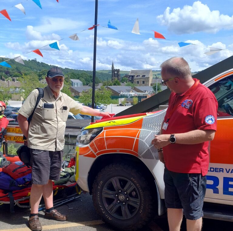 Don and Rob stand outdoors next to a white and orange rescue vehicle, with colorful bunting hanging above against a backdrop of hills and a church tower.