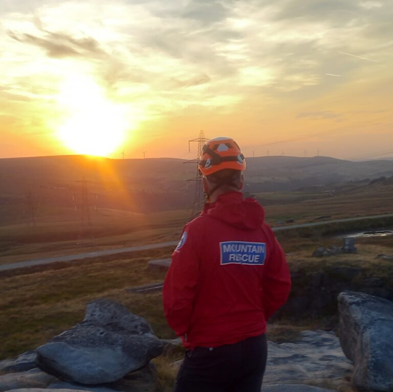 A mountain rescue volunteer in a red jacket and orange helmet stands on a rocky hill, looking out at a scenic sunset over a rolling landscape.